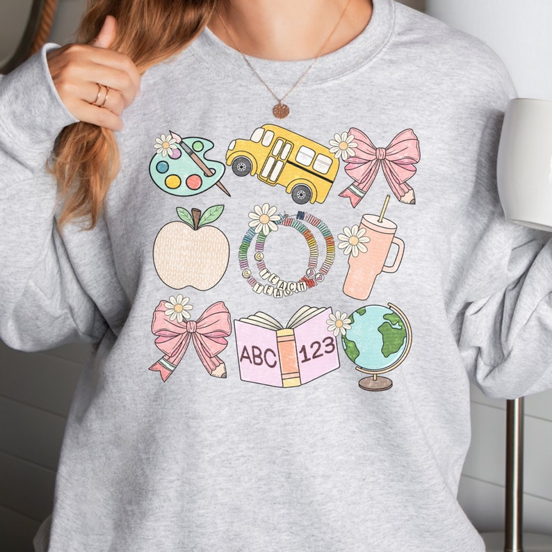 May include: A light gray sweatshirt featuring colorful school-themed illustrations. The design includes a yellow school bus, a pink bow with a pencil, an apple, a book with "ABC 123", a globe, and a paint palette. The sweatshirt has a crew neck.
