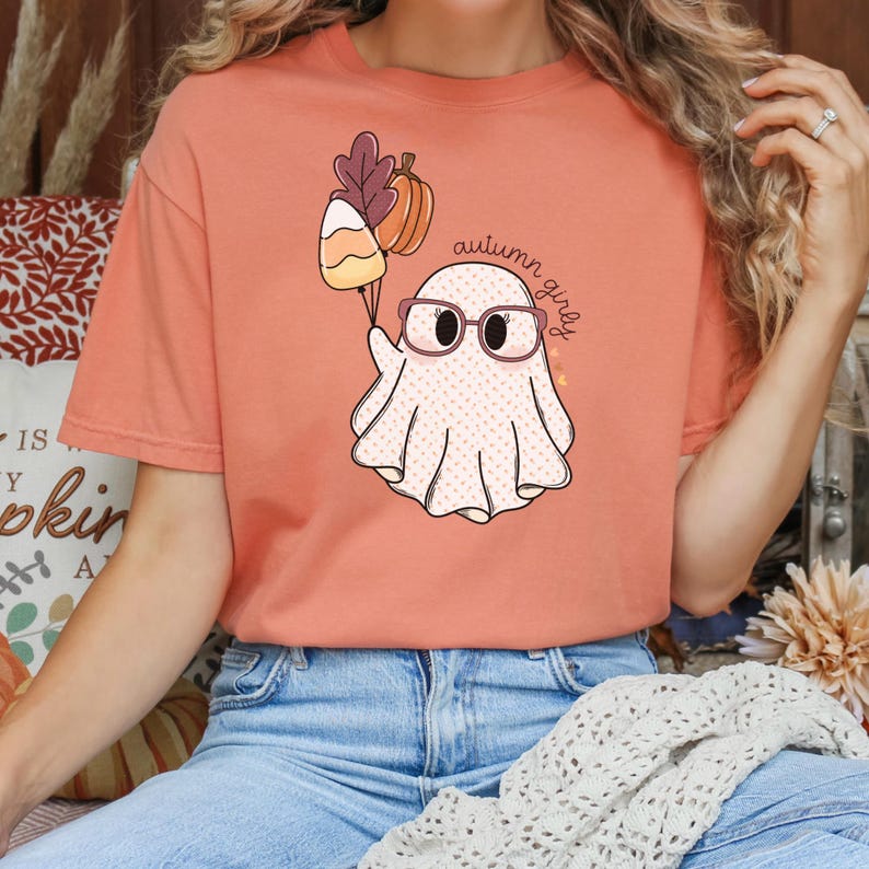 May include: A peach coloured t-shirt with a cartoon ghost wearing glasses and holding a bunch of balloons. The balloons are a candy corn, a leaf, and a small orange pumpkin. The text "autumn girls" is written on the shirt.