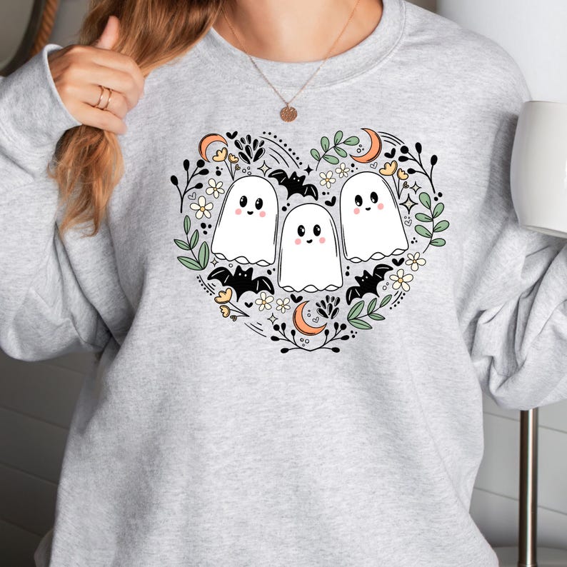 Op de afbeelding: Een grijs sweatshirt met een wit hartvormig ontwerp met drie cartoon-geesten, vleermuizen en bloemen. De geesten hebben glimlachende gezichten en zijn omringd door groene bladeren en witte bloemen.