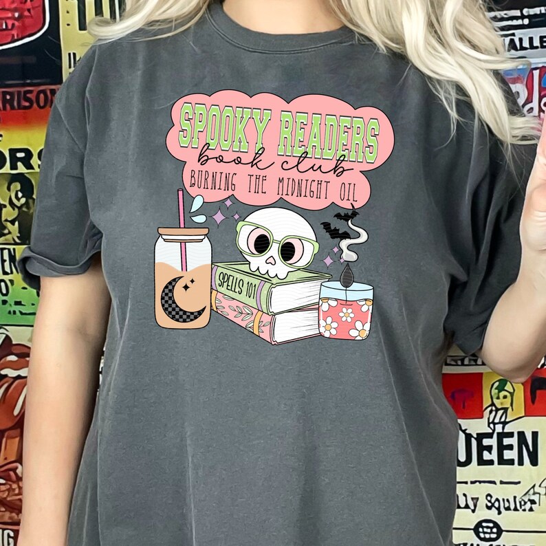 May include: A dark gray t-shirt with a pink and green graphic design that says "Spooky Readers Book Club Burning the Midnight Oil". The graphic includes a skull wearing glasses, a stack of books, a candle, and a glass jar with a moon and stars.