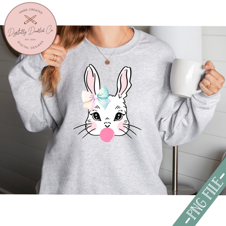 May include: A white bunny with pink cheeks and a pastel rainbow bow is blowing a pink bubble. The bunny is on a gray sweatshirt.