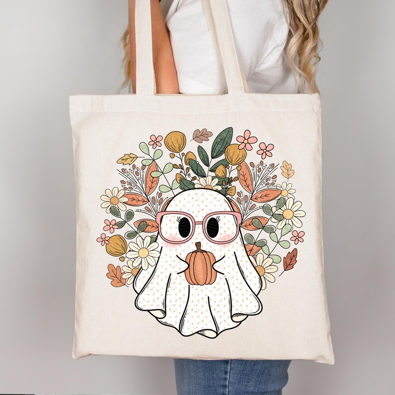 May include: A white tote bag with a cute cartoon ghost wearing pink glasses and holding a pumpkin. The ghost is surrounded by a floral wreath with orange, yellow, and pink flowers.