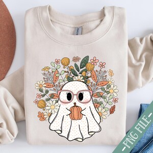 May include: A beige sweatshirt with a cute ghost wearing pink glasses and holding a small orange pumpkin. The ghost is surrounded by a floral design with fall colors like orange, yellow, and green.