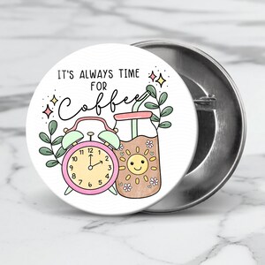 May include: A white button with a pink alarm clock and a brown glass of coffee with a straw. The text on the button reads "It's always time for coffee".