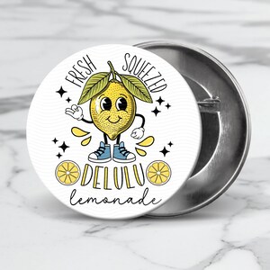 Pu&ograve; includere: Un bottone bianco con un limone di cartone animato che indossa scarpe da ginnastica blu e il testo "Fresh Squeezed Delulu Lemonade".