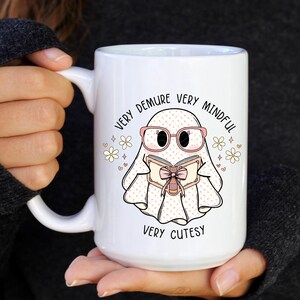 May include: White ceramic mug featuring a pink and white ghost illustration reading a book. The text on the mug reads "Very Demure Very Mindful Very Cutesy".