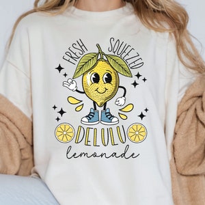 Pu&ograve; includere: Una felpa bianca con una grafica di un limone sorridente con il testo "Fresh Squeezed Delulu Lemonade".