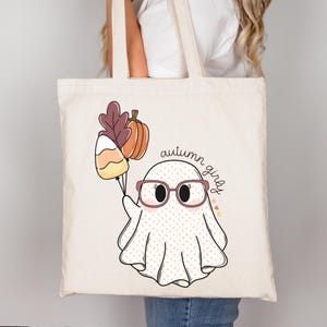 May include: A white tote bag featuring a cute ghost wearing glasses and holding a candy corn and a pumpkin. The text "autumn girly" is written on the bag.