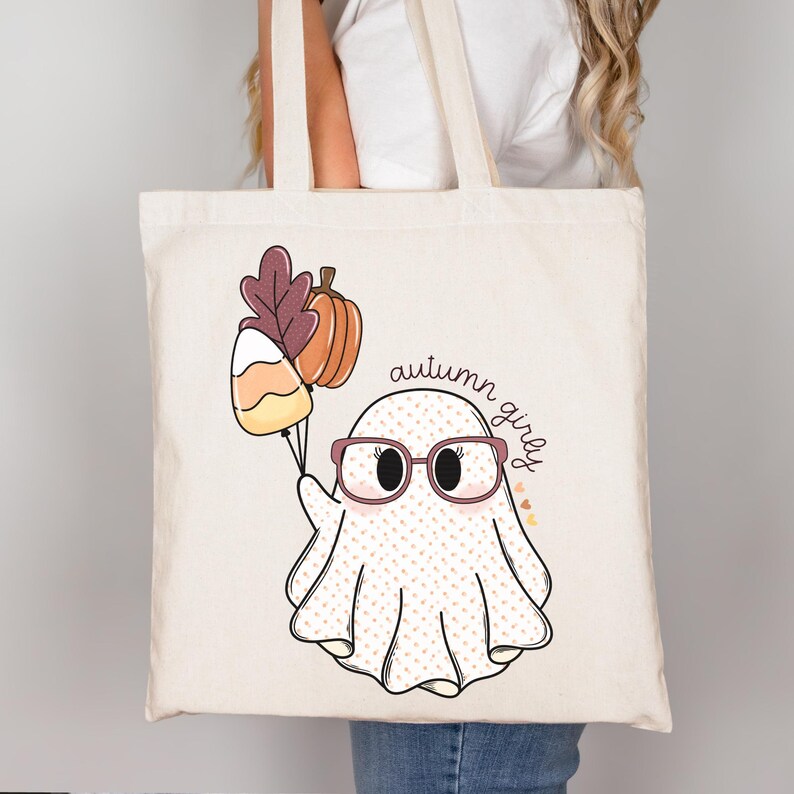 May include: A white tote bag featuring a cute ghost wearing glasses and holding a candy corn and a pumpkin. The text "autumn girly" is written on the bag.