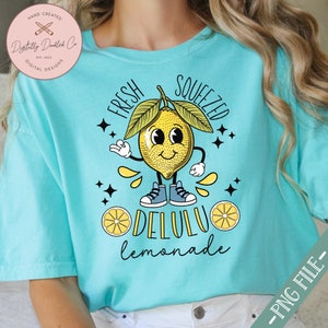 Pu&ograve; includere: Una maglietta turchese con un disegno di limone a cartone animato. Il limone ha un viso sorridente e indossa scarpe da ginnastica. Il testo "FRESH SQUEEZED DELULU lemonade" &egrave; stampato sulla maglietta.