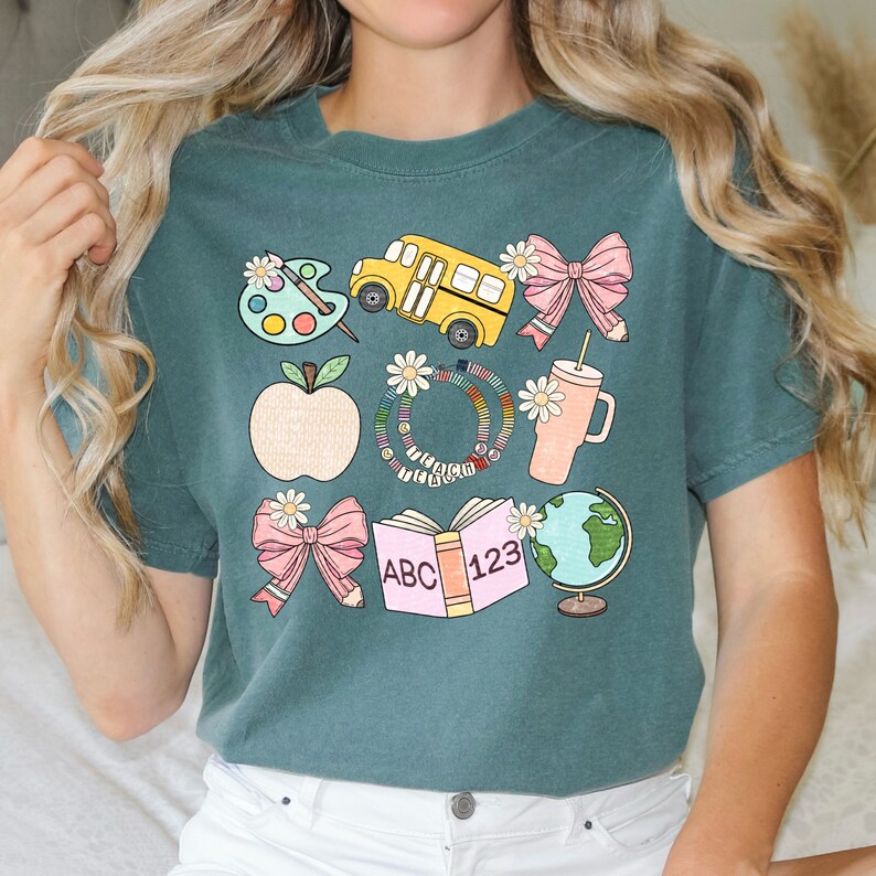 May include: Teal short-sleeve t-shirt with a colorful school-themed graphic. The design includes a yellow school bus, an apple, a palette, a globe, a book with "ABC 123", and daisy accents.