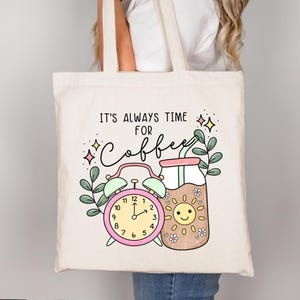 May include: A white canvas tote bag with the text "IT'S ALWAYS TIME FOR COFFEE" in black. The design features a pink alarm clock, a brown glass with a straw, and a yellow sun with a smiling face.