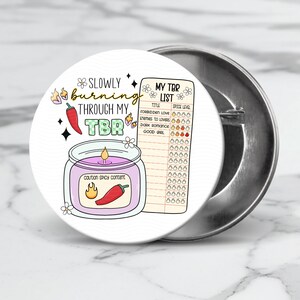 May include: A white button with a colorful illustration of a candle with a red chili pepper and the text "Slowly burning through my TBR".  The button also features a list titled "My TBR List" with a spice level chart.