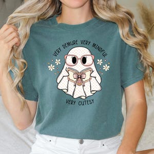 May include: Teal blue t-shirt featuring a white ghost wearing pink glasses and reading a book with a pink bow. The text on the shirt reads "Very Demure Very Mindful Very Cutesy".