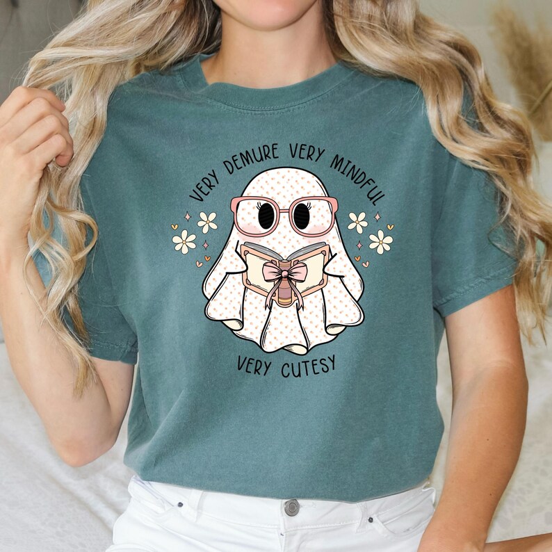 May include: Teal blue t-shirt featuring a white ghost wearing pink glasses and reading a book with a pink bow. The text on the shirt reads "Very Demure Very Mindful Very Cutesy".