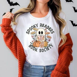 Pode incluir: Camiseta branca com um gr&aacute;fico de um fantasma lendo um livro cercado por flores e ab&oacute;boras. O texto "Spooky Readers Book Society" est&aacute; escrito em um c&iacute;rculo ao redor do fantasma.