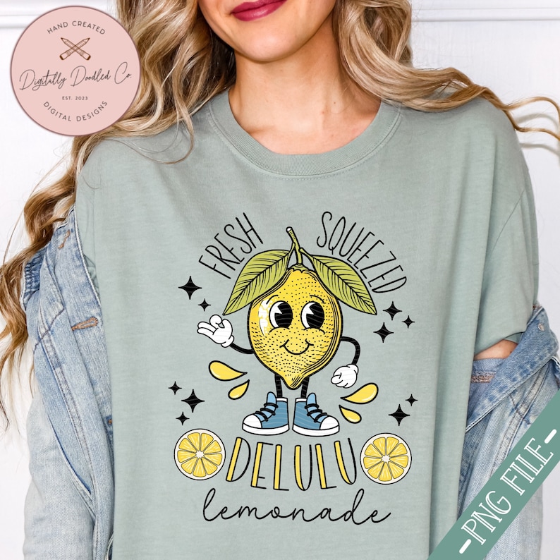 Pu&ograve; includere: Una maglietta verde chiaro con un disegno di limone a cartone animato. Il limone indossa scarpe da ginnastica e ha un viso sorridente. Il testo "Fresh Squeezed DELULU lemonade" &egrave; stampato sulla maglietta.