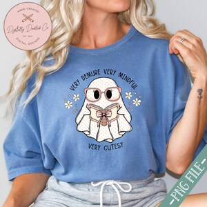 May include: A blue t-shirt featuring a white ghost wearing pink glasses and reading a book. The ghost has the text "Very Demure Very Mindful Very Cutesy" around it.