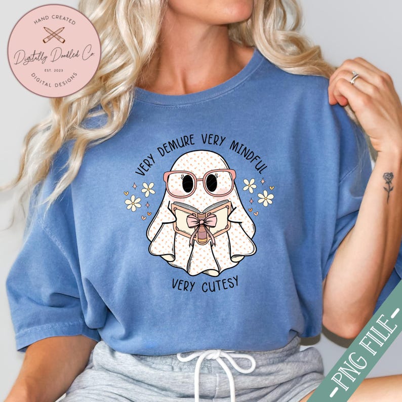 May include: A blue t-shirt featuring a white ghost wearing pink glasses and reading a book. The ghost has the text "Very Demure Very Mindful Very Cutesy" around it.