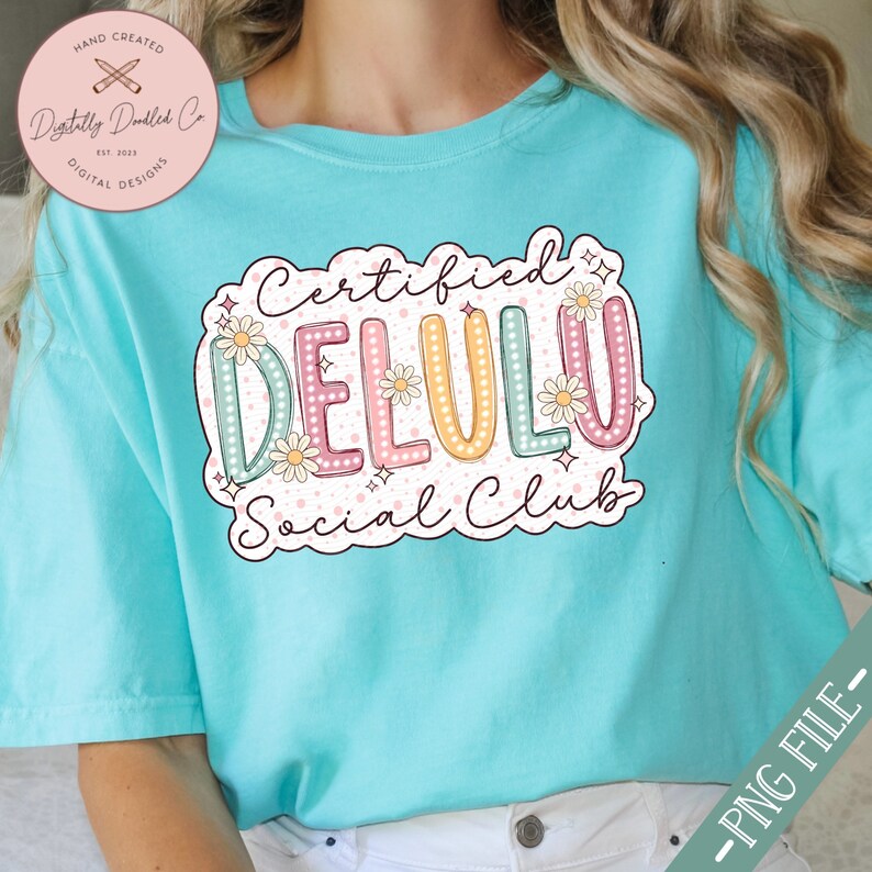 May include: A turquoise t-shirt with a graphic design that says "Certified Delulu Social Club". The graphic is in a colorful, cartoon style with flowers and sparkles.