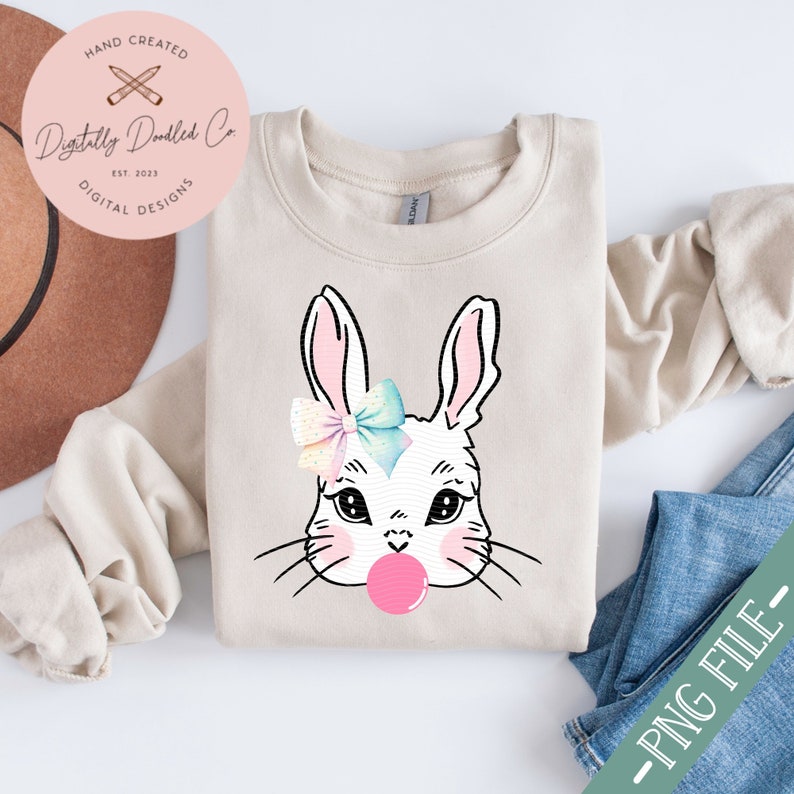 May include: A beige sweatshirt with a white bunny face wearing a pastel bow and blowing a pink bubble. The bunny has large black eyes and a pink nose. The sweatshirt is folded and laid on a white surface with a brown hat and blue jeans.
