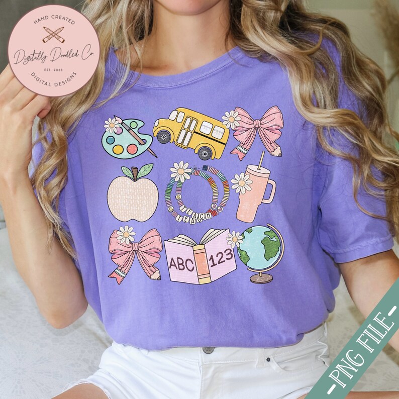 May include: A purple t-shirt featuring school-themed graphics. The design includes a yellow school bus, an apple, a globe, a book with "ABC 123", bows, a palette, a drink cup, and a daisy-adorned bracelet with the word "TEACHER".