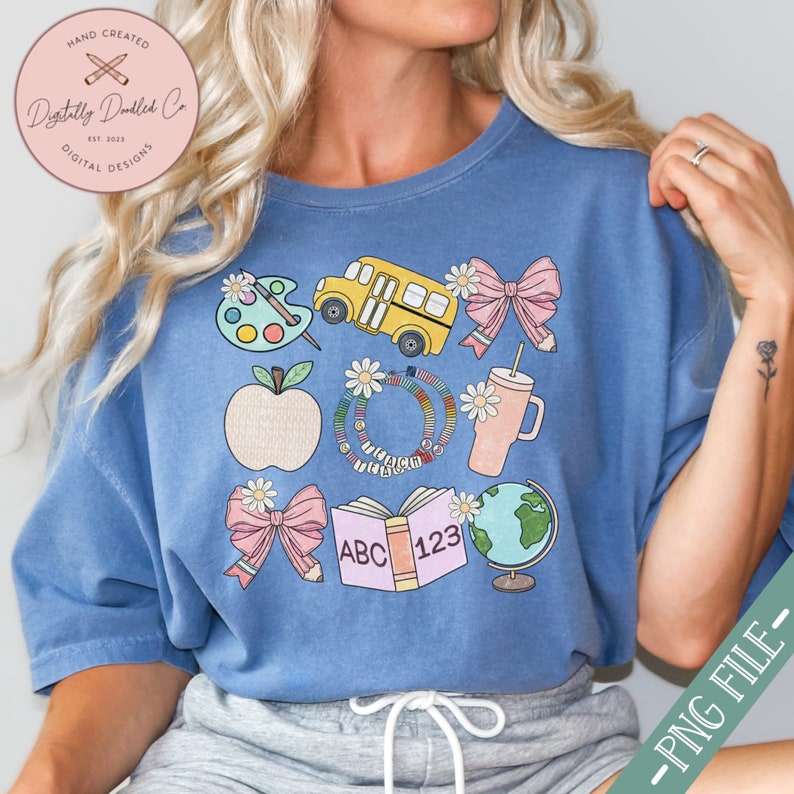 May include: A blue t-shirt featuring a collection of school-themed illustrations. The design includes a yellow school bus, a palette with paint, an apple, bows, a tumbler, a globe, and an open book with "ABC 123". The shirt also has the text "Digitally Doodled Co" and "EST. 2023".