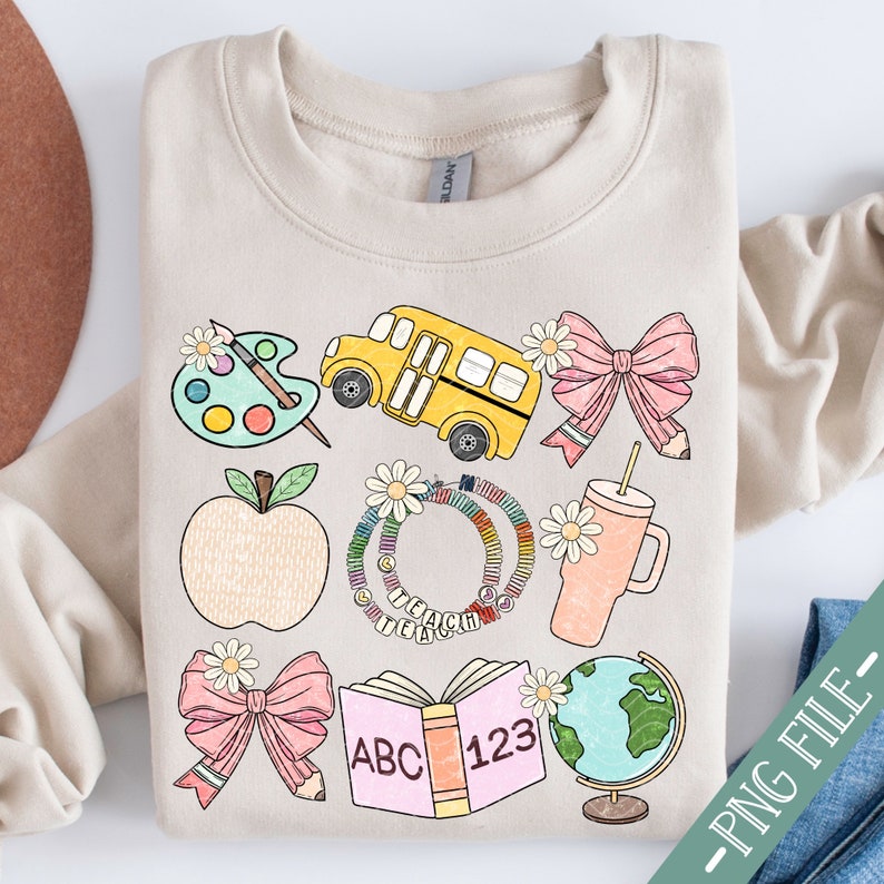 May include: Beige sweatshirt with colorful school-themed graphics. Includes a yellow school bus, apple, paint palette, pink bows, a tumbler, a globe, an open book with "ABC 123", and a daisy-adorned circle with "TEACHER" text.