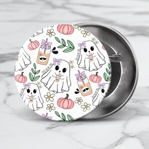 May include: A white button featuring a repeating pattern of ghosts holding jars with the text "Cold Like My Soul" and pumpkins. The background is white with green leaves and white flowers.
