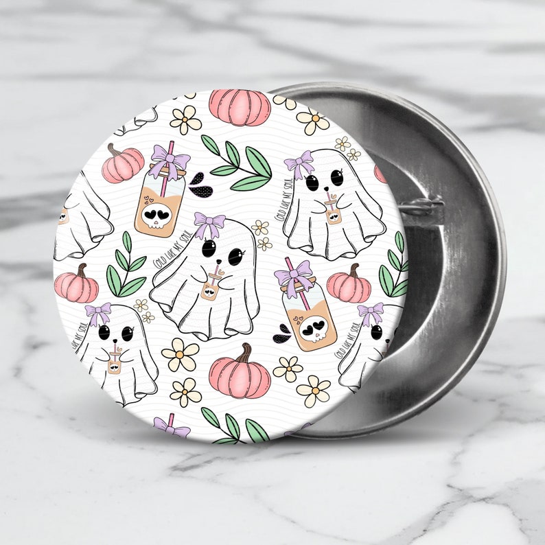 May include: A white button featuring a repeating pattern of ghosts holding jars with the text "Cold Like My Soul" and pumpkins. The background is white with green leaves and white flowers.