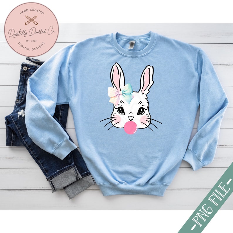 May include: A light blue sweatshirt with a white bunny rabbit wearing a pink bow and blowing a pink bubble. The bunny has black eyes and whiskers.