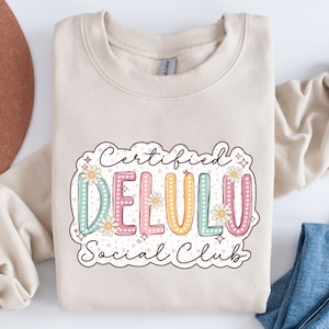 May include: A beige sweatshirt with a graphic design that says "Certified Delulu Social Club". The graphic is in a retro style with colorful letters and flowers.