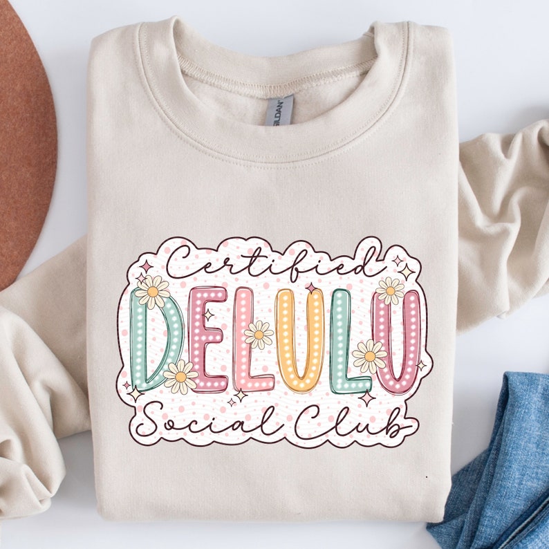 May include: A beige sweatshirt with a graphic design that says "Certified Delulu Social Club". The graphic is in a retro style with colorful letters and flowers.