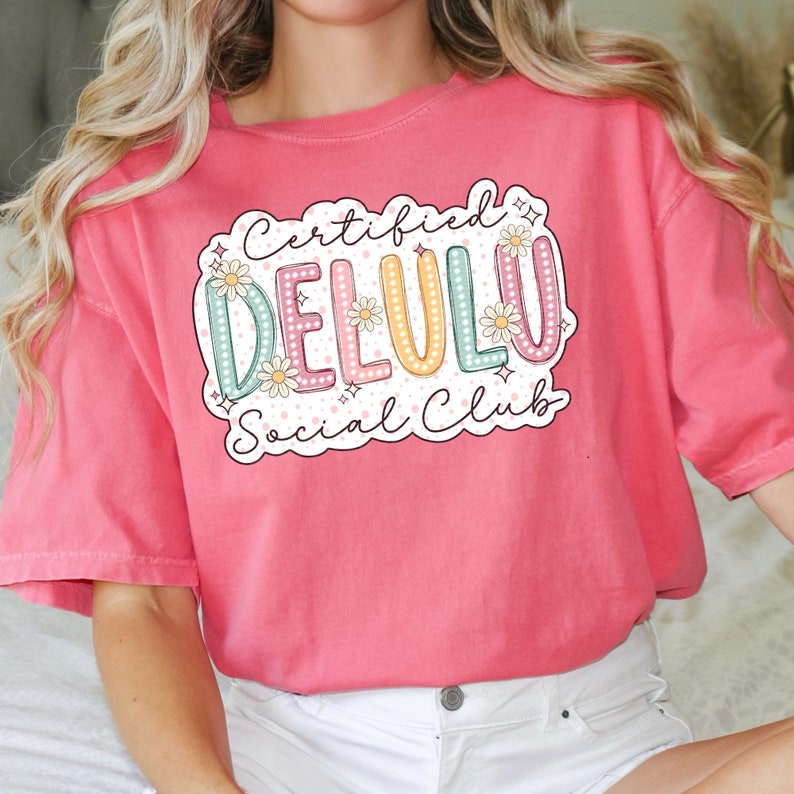 May include: A pink t-shirt with a graphic design that says "Certified Delulu Social Club" in a colorful, retro font with flowers and sparkles.