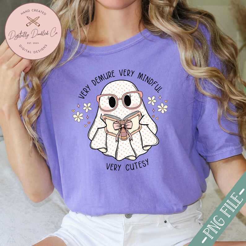 May include: Purple t-shirt featuring a cartoon ghost wearing glasses and reading a book. The text on the shirt reads "Very Demure Very Mindful Very Cutesy".