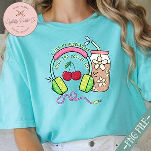 May include: A turquoise blue t-shirt with a graphic design of headphones, cherries, and a glass of iced coffee. The text on the shirt reads "I like my audiobooks spicy and coffee icy."