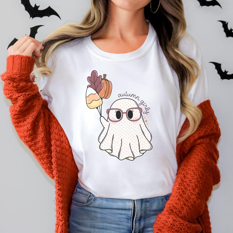 May include: White t-shirt featuring a cartoon ghost wearing glasses and holding a candy corn and a pumpkin. The text "autumn girly" is written on the shirt.