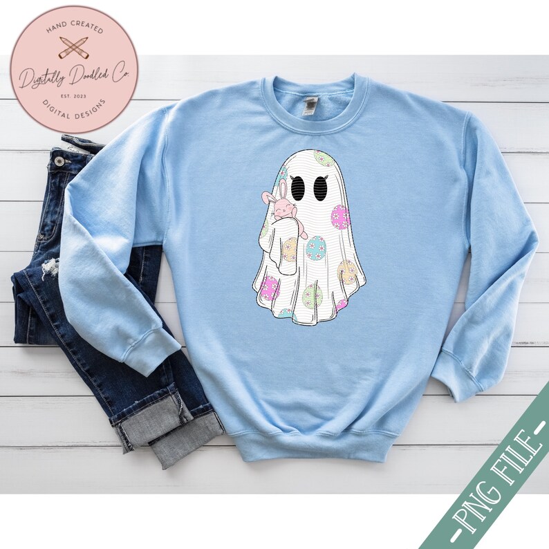 May include: A light blue sweatshirt with a white ghost holding a pink bunny. The ghost is decorated with pastel colored polka dots and floral designs.