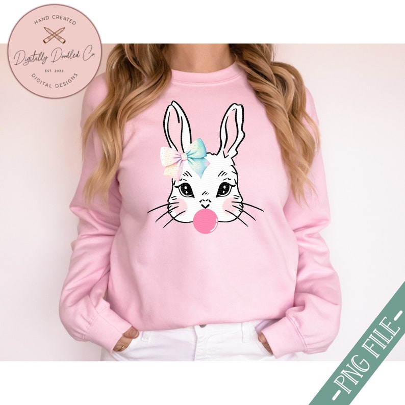 May include: A pink sweatshirt with a white bunny wearing a blue and pink bow and blowing a pink bubble. The bunny has black eyes and a black nose.