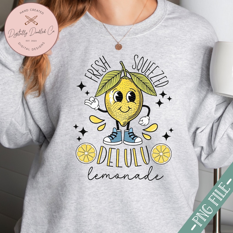 Pu&ograve; includere: Una felpa grigia con un design a limone di cartone animato. Il limone indossa scarpe da ginnastica e ha un'espressione allegra. Il testo "Fresh Squeezed Delulu Lemonade" &egrave; stampato sulla felpa.