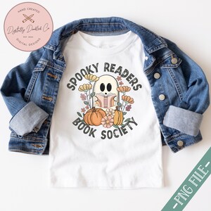 Pode incluir: Camiseta branca com um gr&aacute;fico de um fantasma lendo um livro cercado por flores e o texto "Spooky Readers Book Society".