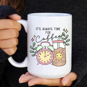 May include: White ceramic mug with a pink alarm clock and a glass of coffee with a sun face. The mug has the text "It's always time for coffee" in black.