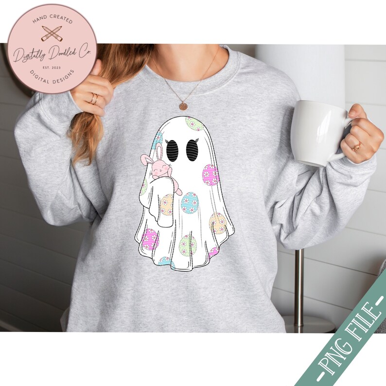 May include: A gray sweatshirt with a white ghost holding a pink bunny. The ghost is decorated with pastel colored polka dots. The text "Digitally Doodled Co. Est. 2023 Digital Designs" is on a brown tag.