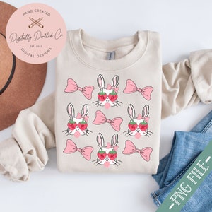 May include: A beige sweatshirt with a repeating pattern of white bunnies wearing pink bows and strawberry sunglasses. Each bunny is blowing a pink bubble. The text "Digitally Doodled Co. Est. 2023 Digital Designs" is on a white circle in the top left corner.