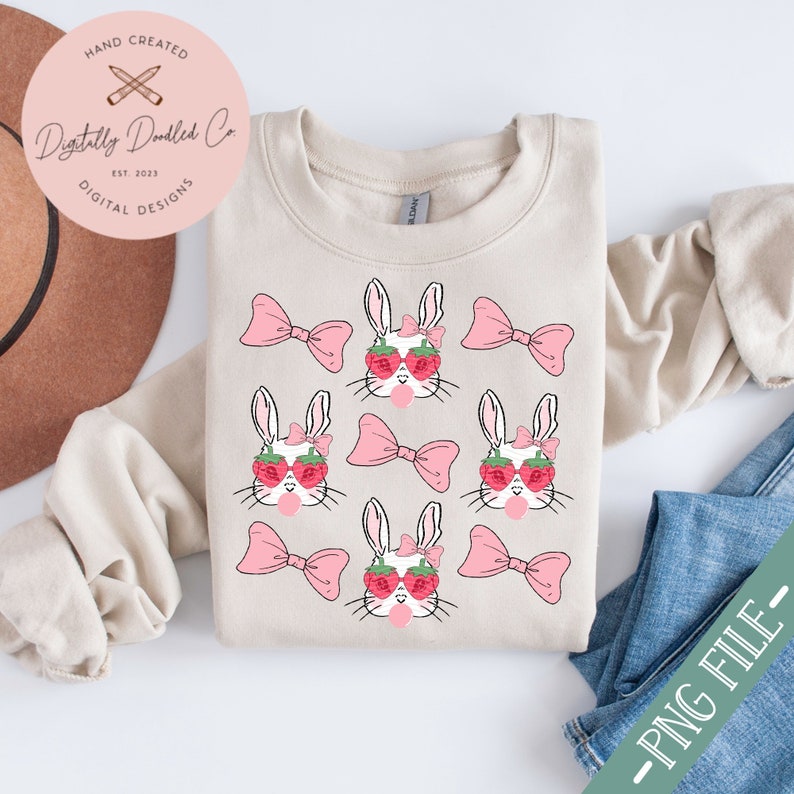 May include: A beige sweatshirt with a repeating pattern of white bunnies wearing pink bows and strawberry sunglasses. Each bunny is blowing a pink bubble. The text "Digitally Doodled Co. Est. 2023 Digital Designs" is on a white circle in the top left corner.