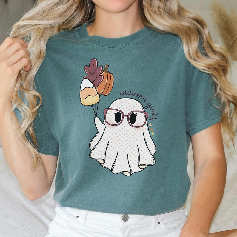 May include: Teal blue t-shirt featuring a white ghost wearing glasses and holding a candy corn and a pumpkin. The text "autumn girly" is written on the shirt.