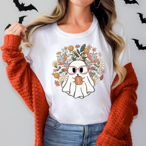 May include: White t-shirt with a cute cartoon ghost wearing glasses and holding a pumpkin. The ghost is surrounded by fall-colored flowers and leaves.