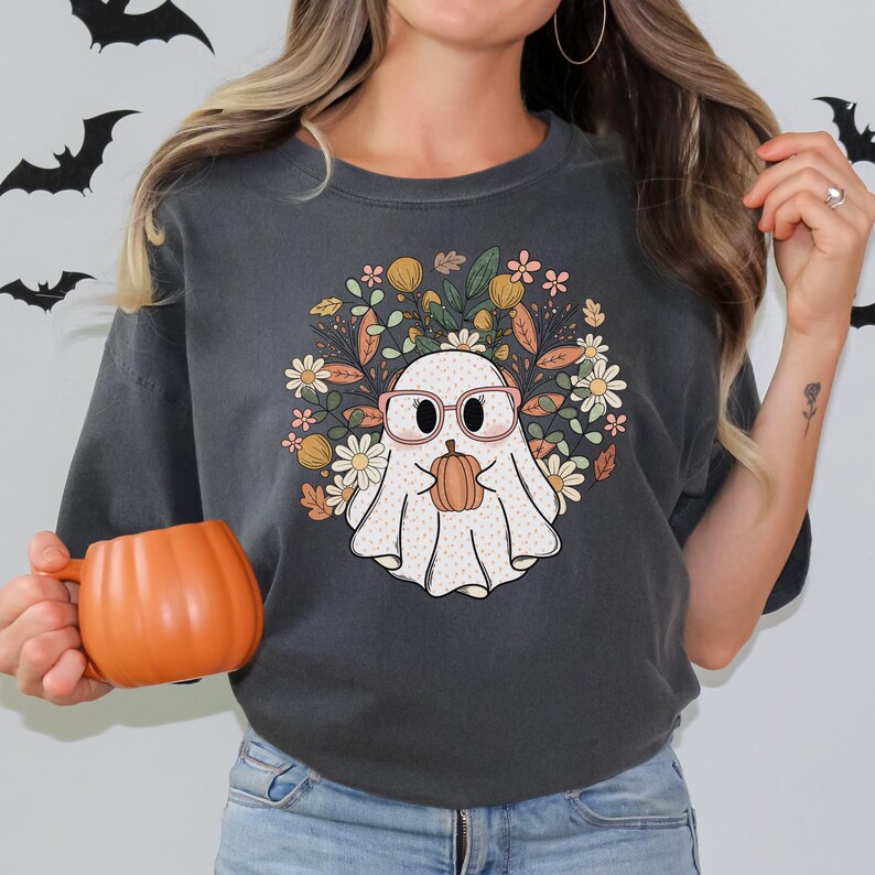 May include: A dark gray t-shirt with a white ghost wearing pink glasses and holding a small orange pumpkin. The ghost is surrounded by a floral design with pink, yellow, and green flowers.