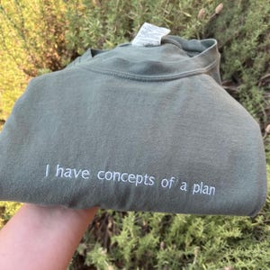 May include: A green t-shirt with the text "I have concepts of a plan" embroidered in white.