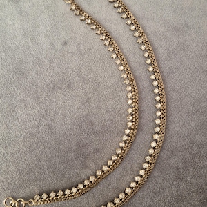 May include: Two gold-coloured anklets with a delicate chain design. Each anklet features a row of small, sparkling clear crystals. The anklets have a clasp closure and are displayed on a grey surface.